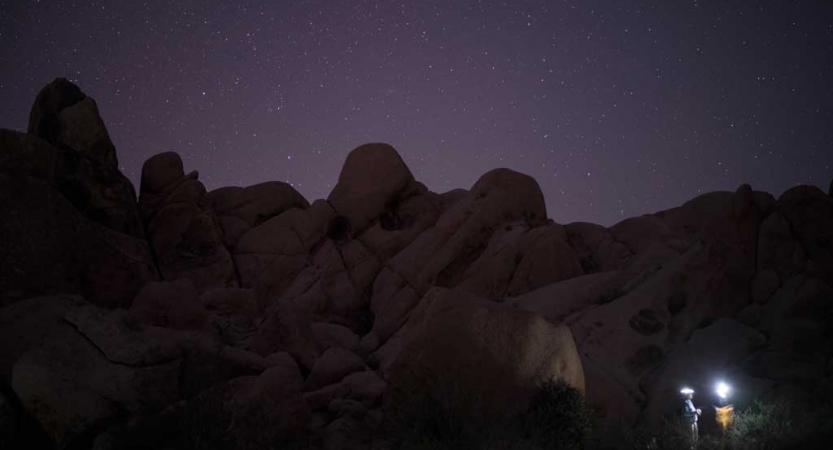 the headlamps of two people are illuminated in the bottom right of the photo. Behind them, a rocky ridge outlines the purple sky above.
